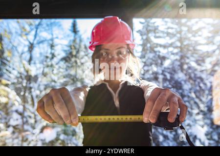 Selective focus on girl's hands showing tape measure, smiling architect woman wearing a pink helmet and protective glasses with snowy trees background Stock Photo