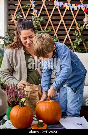 Two people (mother and son) carving pumpkins at a decorated table in Autumn, suggesting a festive, autumnal activity. Woman and child making a pumpkin Stock Photo