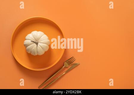 Autumn and Thanksgiving Day table setting with white small pumpkin, orange empty plate with golden cutlery on orange background. Concept catering. Top Stock Photo
