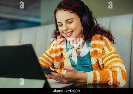A smiling female college student wearing headphones, studying online with a smartphone and laptop at a cozy cafe. Emphasizes modern education, technol Stock Photo