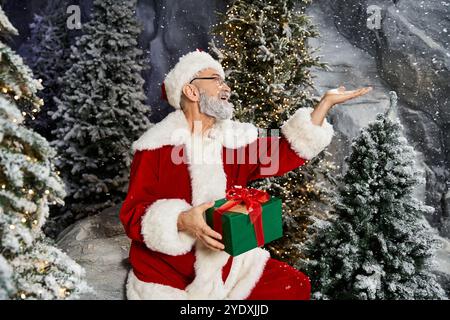 A cheerful Santa holds a wrapped gift surrounded by snow covered trees. Stock Photo