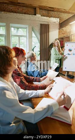 Project leader outlines strategic plan on whiteboard, with team members actively engaged in discussion in modern meeting room. Stock Photo