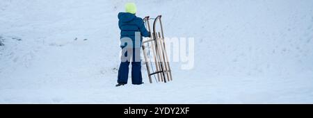 A child dressed in a blue jacket and neon beanie stands on a snow-covered hill, holding a wooden sled, ready for a winter ride. Stock Photo
