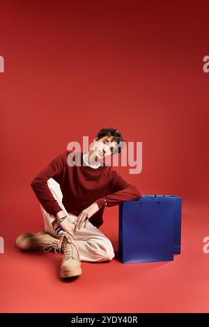 A handsome young man with curly hair sits casually, showcasing shopping bags amidst a bold backdrop. Stock Photo