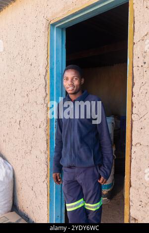 portrait of a village young African man , construction worker standing in the door frame of the mud house , cement bags and paint drums in the backgro Stock Photo
