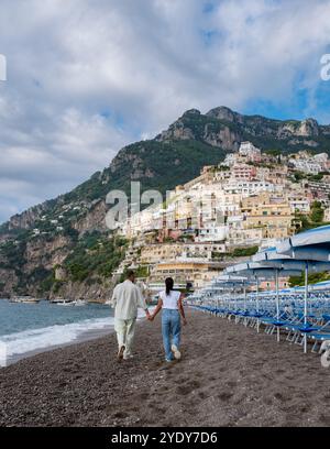 A couple walks hand in hand along the pebbled beach of the Amalfi Coast, surrounded by vibrant cliffside homes. Stock Photo