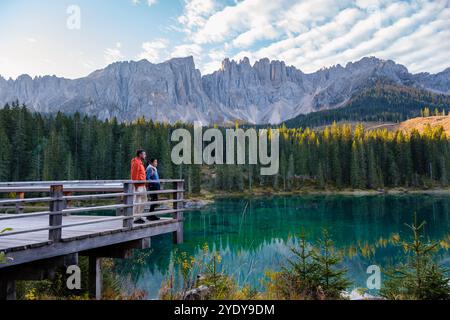 In the heart of the Dolomites, two hikers stand on a wooden deck, captivated by the turquoise lake and impressive mountain peaks. Lake Carezza or Karersee Dolomites in Italy. Stock Photo