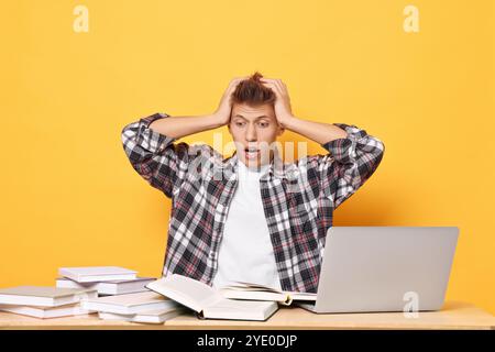 Emotional student with books and laptop having stress before exam at table against yellow background Stock Photo