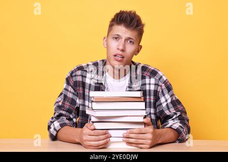 Young student with stack of books having stress before exam at wooden table against yellow background Stock Photo