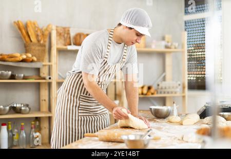 Portrait of confident professional hispanic young baker in apron kneading and working with raw yeasted dough in bakery Stock Photo