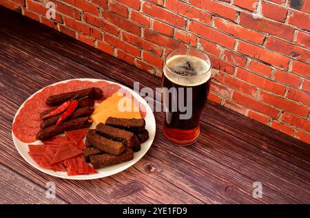 A glass of semi-dark beer and a large plate with an assortment of meat snacks, cheese and rye croutons on a dark wooden table. Close-up. Stock Photo