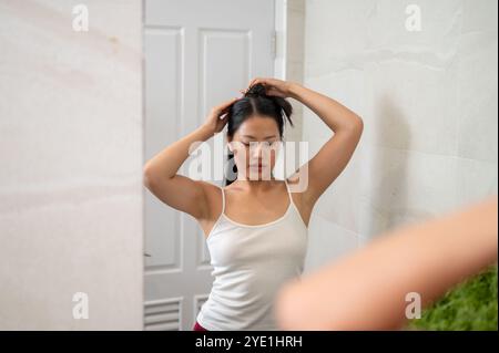 A beautiful Asian woman in pajamas is tying her hair in front of the mirror in the bathroom as she gets ready after her shower. Stock Photo
