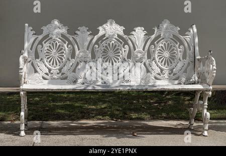 One empty bench made of white iron stands on city park. Sunlight on backrest surface of Victorian pattern white cast iron garden bench in a shady area Stock Photo