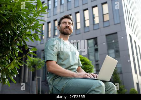 Medical worker man in uniform typing on laptop surfing internet outdoor near hospital Stock Photo