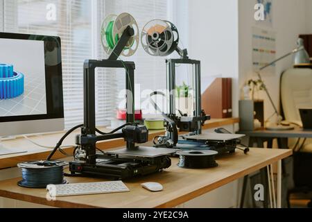 Dual 3D printers operating on wooden desk beside computer displaying object model. Office environment featuring various tech gadgets and decor pieces Stock Photo