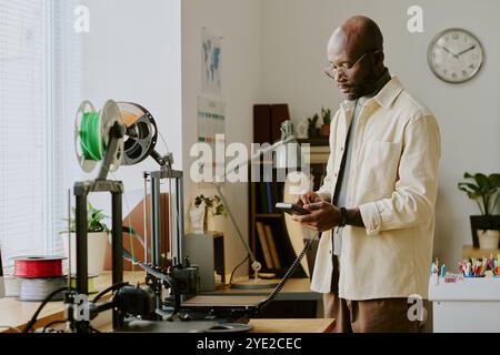 Man reading smartphone while standing in office with 3D printer and various gadgets around desk. Books and plants decorating the background Stock Photo