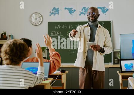 Teacher standing in front of classroom engaging with students while explaining subjects. Maps and technology are visible, indicating a contemporary learning environment Stock Photo