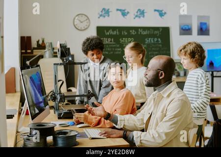 Teacher engaging students while conducting 3D modeling lesson. Group of diverse students focused on classroom setting with computer and 3D printer equipment present Stock Photo