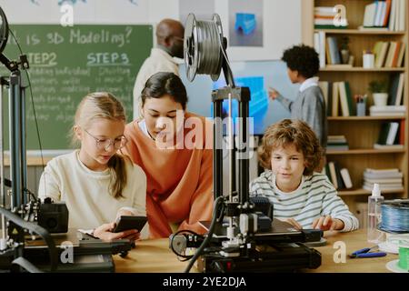 Teacher guiding students through 3D printing process in technology class, explaining equipment usage and modeling techniques. Board in background with steps written on it Stock Photo