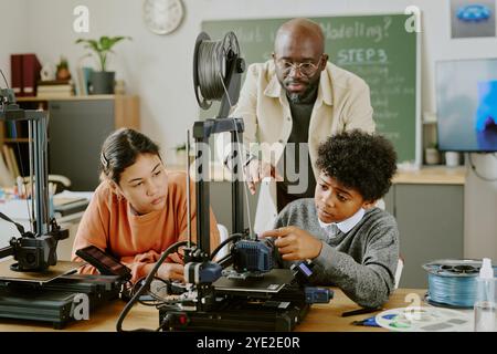 Teacher guiding two students in using 3D printing technology in classroom equipped with 3D printers and computers, providing hands-on educational experience Stock Photo