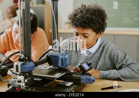 Boy working with 3D printer in classroom, carefully observing process. Engaging in STEM education, collaborating with classmates, wearing school uniform Stock Photo