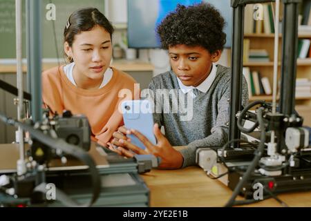 Two students observing a 3D printer's progress in a classroom setting involving technology education. Close-up view of their focused expressions showing concentration around printer setup equipment Stock Photo