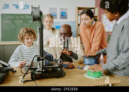 Diverse group of students gathered around a teacher explaining 3D modeling using a 3D printer in classroom setting with educational posters on walls Stock Photo