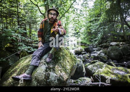 A young man sitting on the stone in the forest. Funny looking male looking at the camera and showing calling hand gesture, come here or whats up Stock Photo