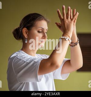 Close-up portrait of woman practicing kundalini yoga, meditating in room and doing lotus mudra with raised hands at face level Stock Photo