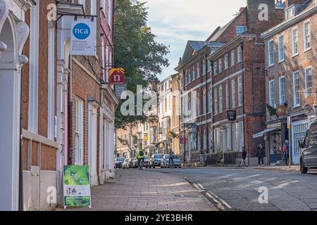 A section of an old cobbled street.  Shops fronts line the pavement and people and cyclist are on the street. Sunlight cut across the road. Stock Photo