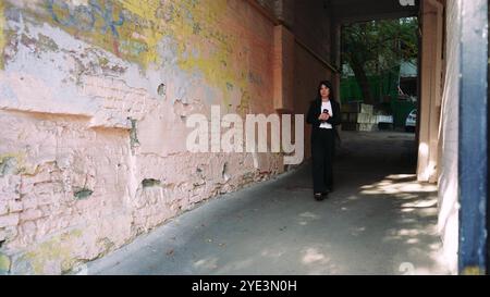 Confidently striding through an archway, a woman in a black suit is framed by the facade of a house in need of repair and shifting shadows conveying Stock Photo