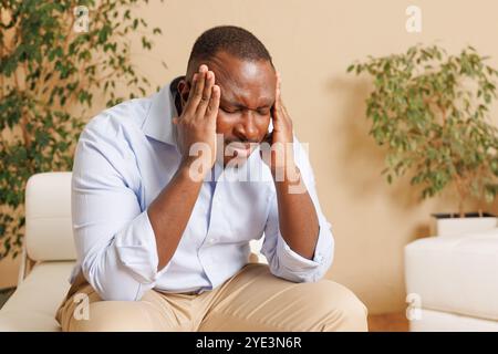 A man sitting on a couch, holding his head with both hands, showing signs of stress or headache. He is wearing a light blue shirt and beige pants Stock Photo