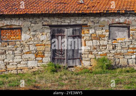 Old stone barn with weathered wooden doors, red tile roof, rustic countryside architecture, vintage farm storage, history and decay in rural setting Stock Photo