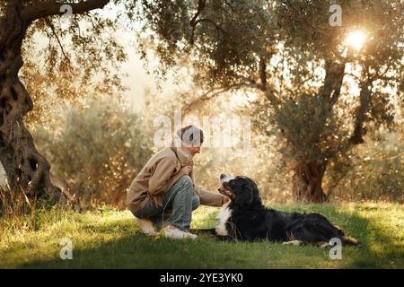 A woman sits peacefully with her Bernese Mountain Dog in a quiet, sun-dappled forest clearing. Stock Photo
