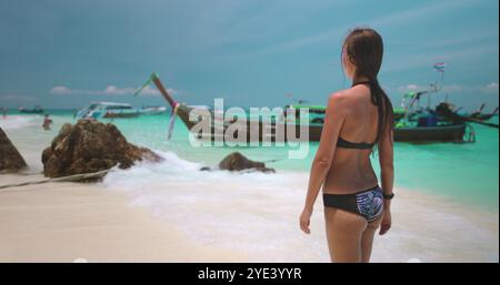 Young woman is enjoying a sunny day on a tropical beach, looking at the horizon where traditional thai longtail boats are moored in the turquoise water. 4K slow motion video Stock Photo