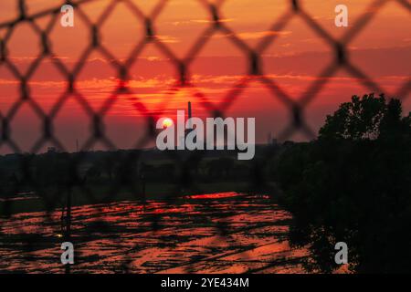 The sun and the factory seen in silhouette against the chain-link fence in the morning Stock Photo