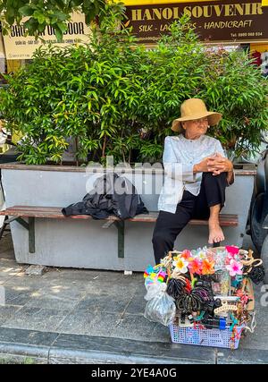 Hanoi, Vietnam: daily Vietnamese life, an elderly woman seated barefoot on a bench selling hair accessories in the streets of the capital city Stock Photo