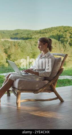 Woman sitting in a chair outdoors, using a laptop to work remotely, surrounded by nature and greenery, enjoying a peaceful environment. Work-life balance Stock Photo