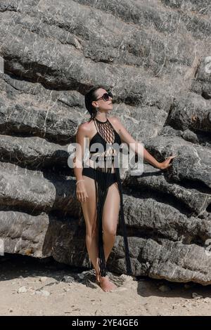 At a picturesque beach, a woman confidently poses beside striking rock formations while wearing a fashionable black swimsuit and stylish sunglasses. Stock Photo
