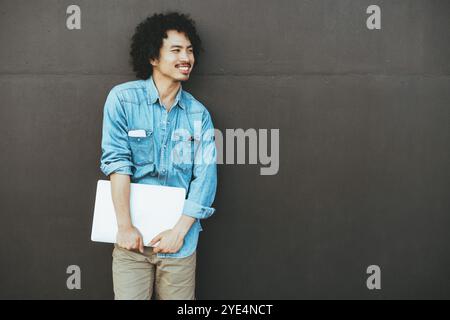Young Filipino man with curly hair, wearing a blue denim shirt, beige pants, holding a white laptop, smiling, looking to the side, standing against a Stock Photo