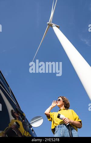Woman with curve hairs wearing yellow shirt and sunglasses stands next to charging electric car. Wind turbine in the background.. Stock Photo