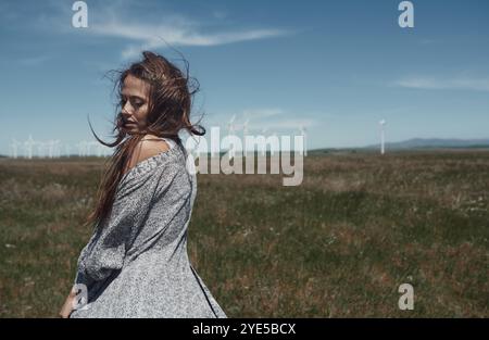 A solitary woman stands in a vast field, her back partly exposed, with wind turbines lining the horizon under a clear blue sky Stock Photo