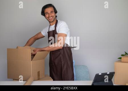 Man in apron happily packing a cardboard box at a table, surrounded by packing materials smiling at camera Stock Photo