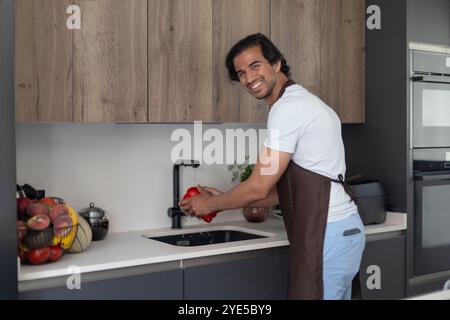 Man in apron washing a red bell pepper in the kitchen, smiling at the camera. Stock Photo