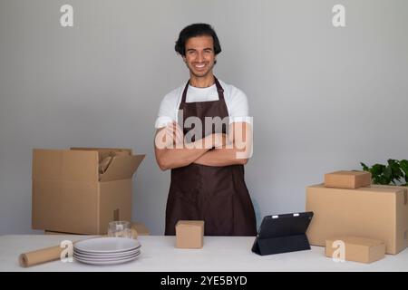 Smiling man wearing a brown apron, standing with arms crossed in a home kitchen with boxes. Stock Photo