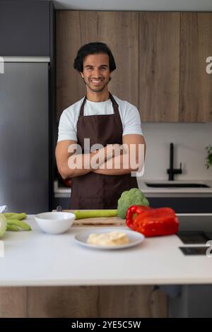 Chef standing confidently with folded arms in a contemporary kitchen, vegetables on counter. Stock Photo
