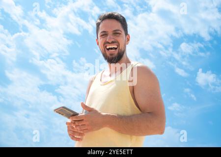 Front image of Young adult Laughing on beach with cell phone looking camera and sunny blue sky Stock Photo