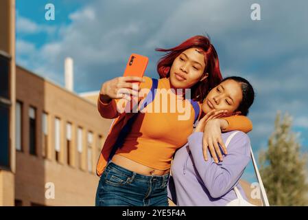 Two young latin women, one with red hair and another with black hair, are seen enjoying a sunny day on a college campus while taking a selfie with a s Stock Photo