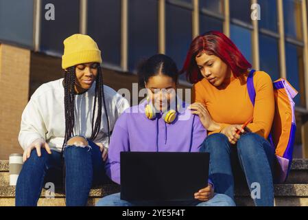 Three young college students, engaging with a laptop while sitting on the campus stairs, reflecting a moment of academic collaboration and youthful ca Stock Photo