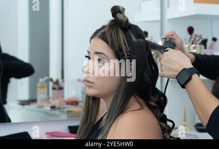 A woman with long hair is getting her hair done by a stylist. The stylist is using a curling iron to style the woman's hair. The woman is relaxed and Stock Photo